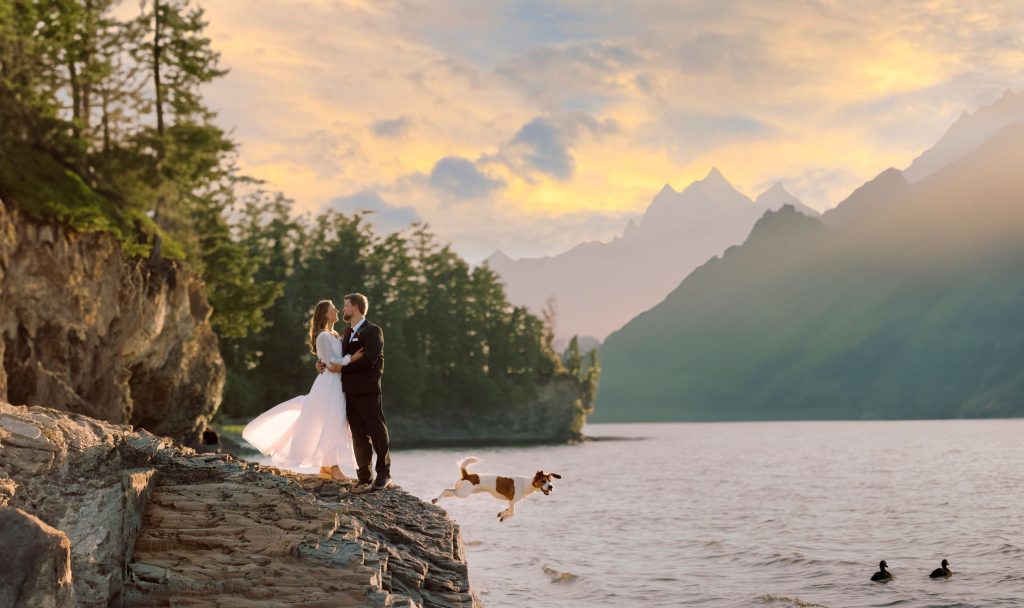 Bride and groom standing on a rocky lakeside cliff during a wedding photoshoot Vermont at sunset, surrounded by mountains, water, and natural scenery.